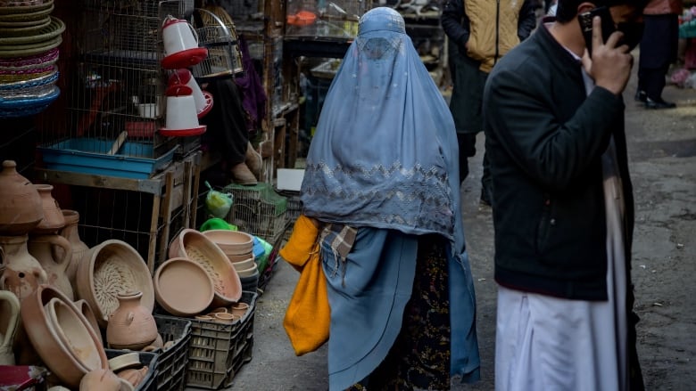 A woman in a blue burqa, covering her head and body, walks in a market holding a yellow bag in her hand. 