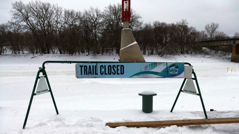 A barricade blocks access to stairs that lead down to Winnipeg's Assiniboine River. A sign reads: Trail closed.