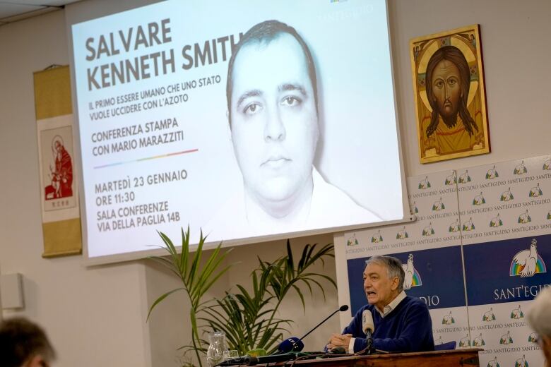 The image of a man is projected on an overhead screen as a man underneath speaks while seated at a desk. 