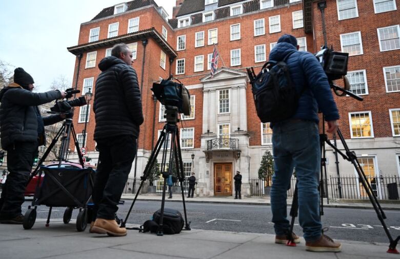 Three people stand on a sidewalk behind media cameras pointed at the front door of a multi-storey red brick building with two police officers outside a door.