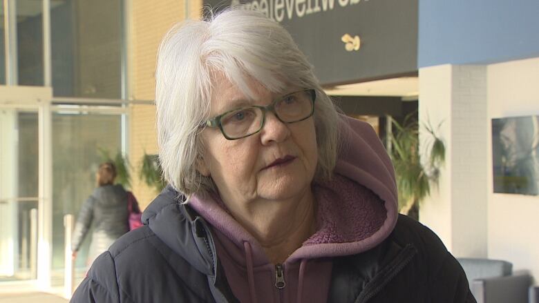 Woman with short white hair and glasses, dressed in parka, inside Credit Union Place lobby. 