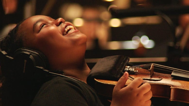 A little girl with headphones tosses her head back and smiles brightly while holding a violin. 