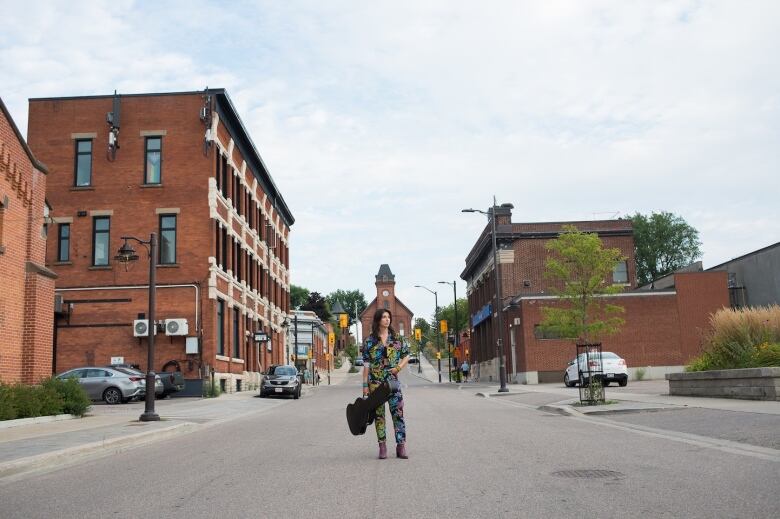 A woman holding a guitar stands in the middle of a small-town main street.