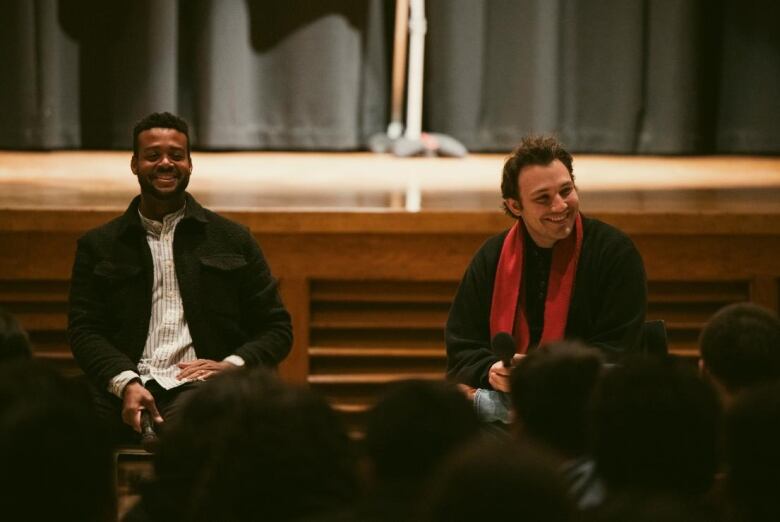 Two men sit side by side, smiling, in front of an audience.