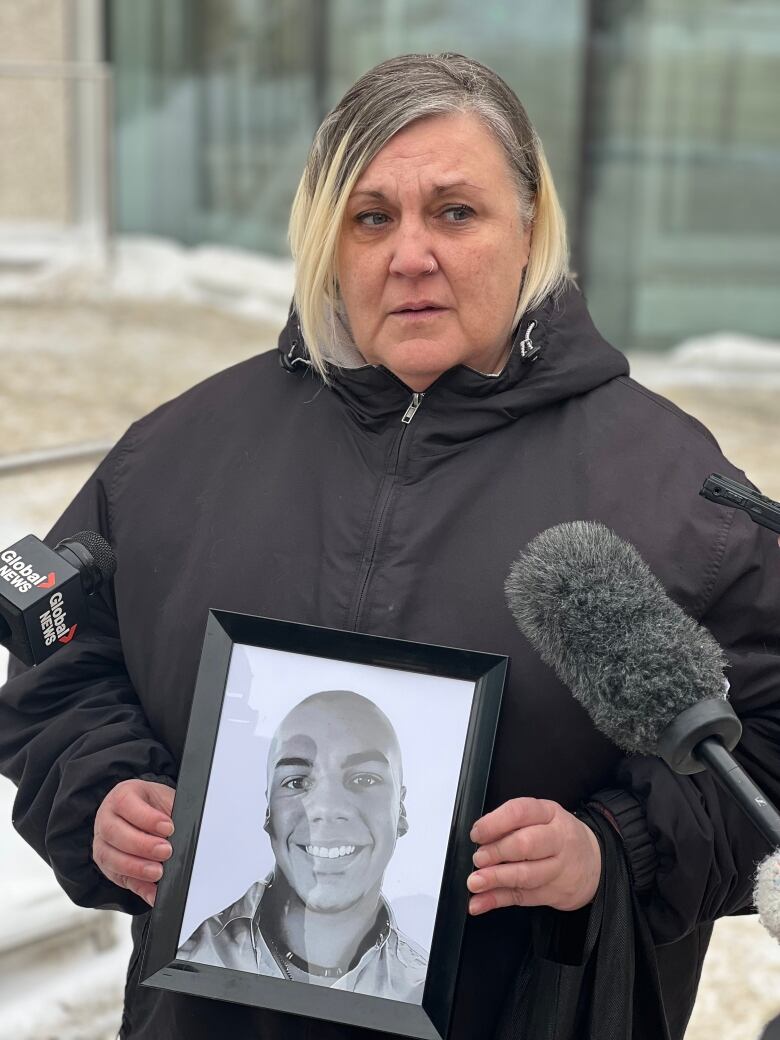 A woman holds a framed photograph of her son as she talks to reporters outside a building.