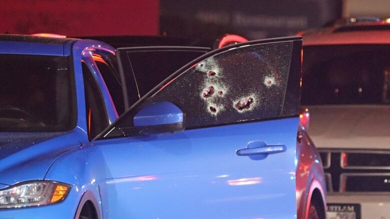 A blue vehicle is pictured with it's driver-side door open, revealing bullet holes in the window.