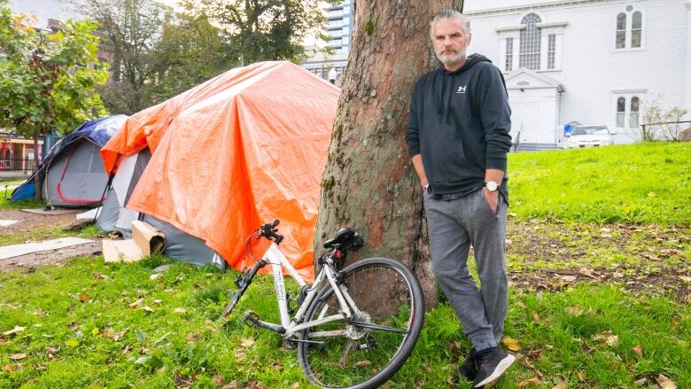 A man leans on a tree next to a bicycle in Halifax's Grand Parade. Behind him are two tents. 