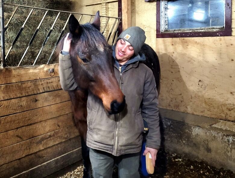 A brown coloured horse leans his head on Jayce Mercer's shoulder in a stable stall. 