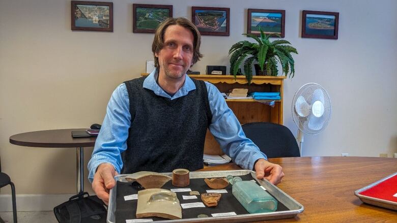 A man sits in front of a tray full of pieces of old ceramic and glass objects.