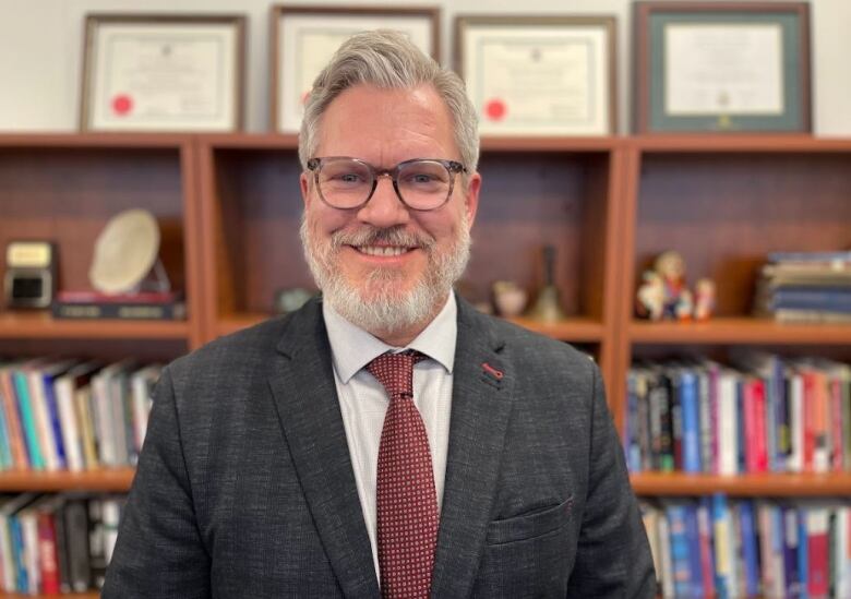 Gray-haired man with beard and glasses poses for photo in front of book shelf.