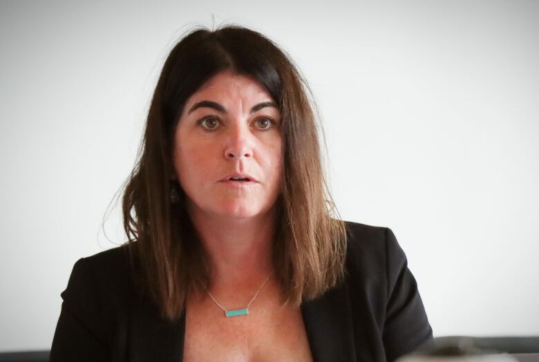 A woman with long brown hair wearing a dark suit jacket speaks to someone off camera.