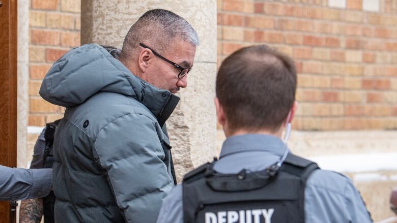 A man a winter jacket is escorted out of a courthouse by a sheriff. 