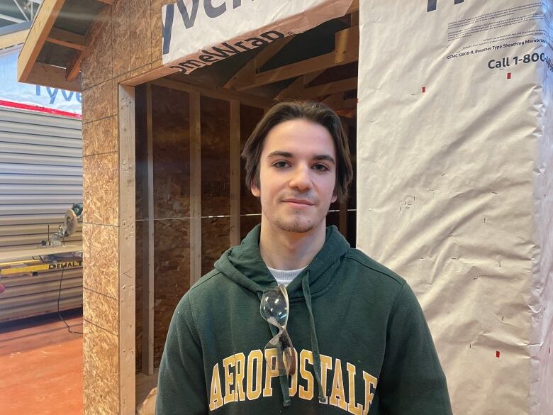 Brayden Doucette poses for a photo. He is wearing a green hoodie with safety glasses hanging off. His hair is brown and he behind him is an unfinished shed. 