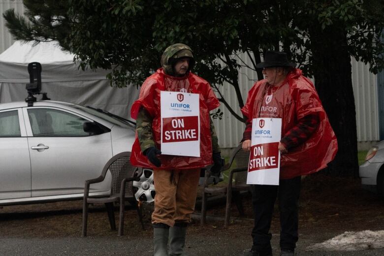 Two men wearing red ponchos and holding sandwich boards reading 'On Strike' stand in the rainy outdoors.