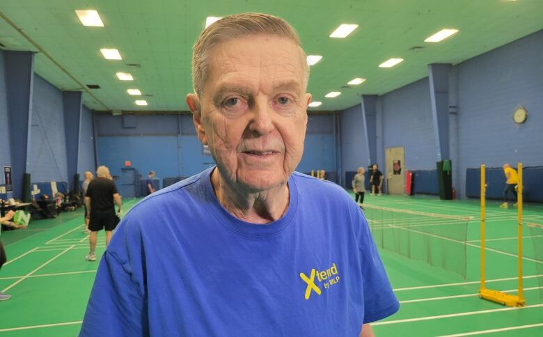 An older gentleman in a blue t-shirt stands on a Pickleball court.