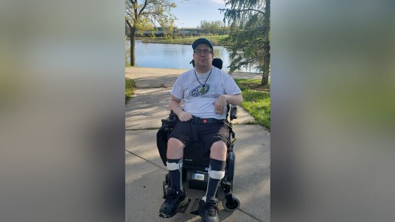 A man in a white T-shirt sits in a wheelchair outside on a summer day.