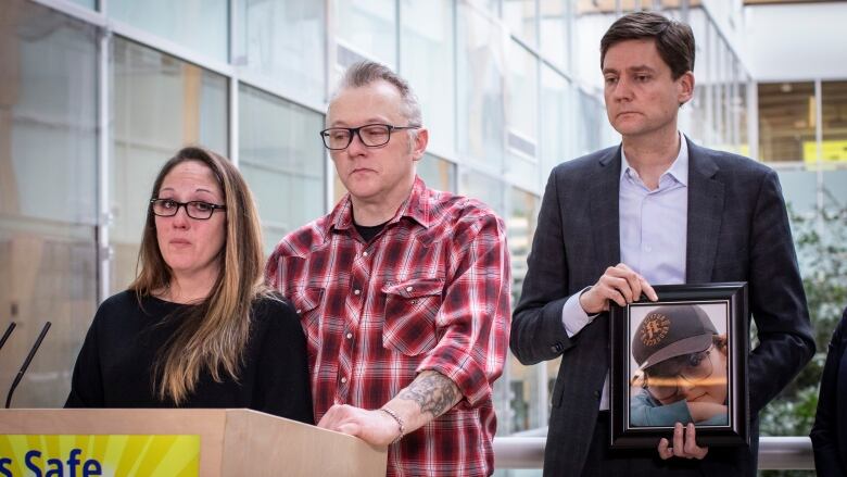 A white man and woman weep at a podium, while a white man behind them holds a picture of a young boy.