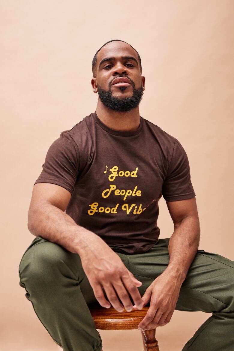 A Black man sits on a stool and looks at the camera.