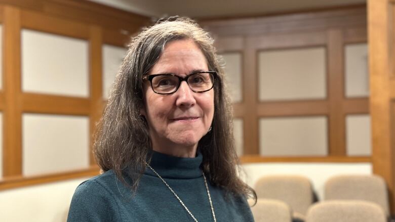 A woman with long grey hair and eyeglasses stands in a wood-paneled room.