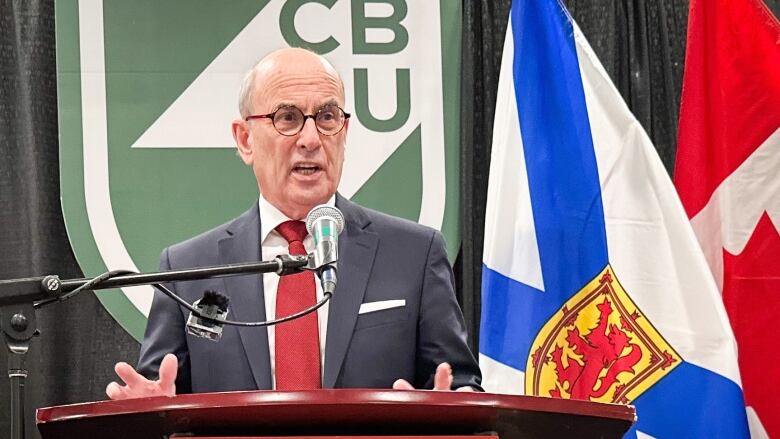 A balding man with round eyeglasses, navy suit, white shirt and red tie speaks at a podium and gestures with his hands, in front of a large green Cape Breton University logo.