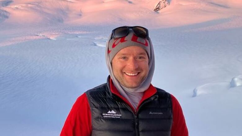 A man with a black jacket on and head coverings stand in snow with snow-capped mountains in the background.