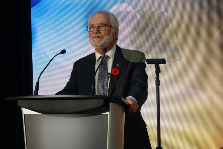 An older man with white hair and beard, wearing glasses and a suit speaks at a podium. Behind him is an LED screen in bright colours.