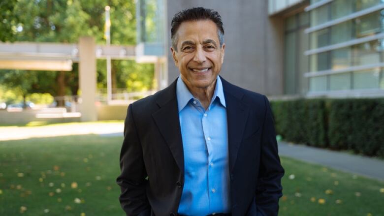 A man with short dark hair, a light blue collared shirt and dark blazer smiles outside with greenery and a grey building in the background.