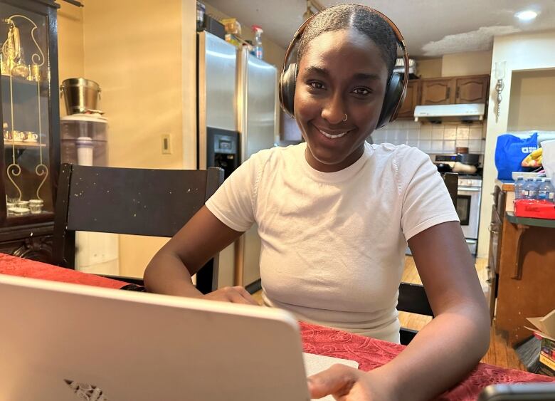 A woman in her 20s sits in front of a laptop at a kitchen table with headphones on.