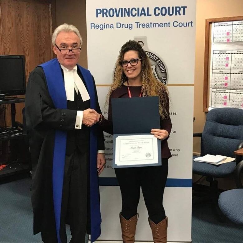 person wearing dark clothing with glasses is shaking hands with person in robe, while receiving a diploma.