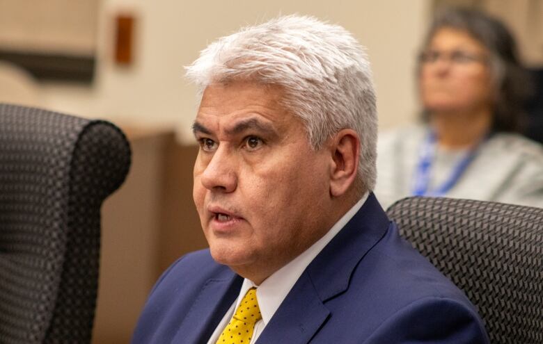 A close-up of a man sitting in a chair. He has white hair and is wearing a blue suit jacket with a white shirt and gold tie.