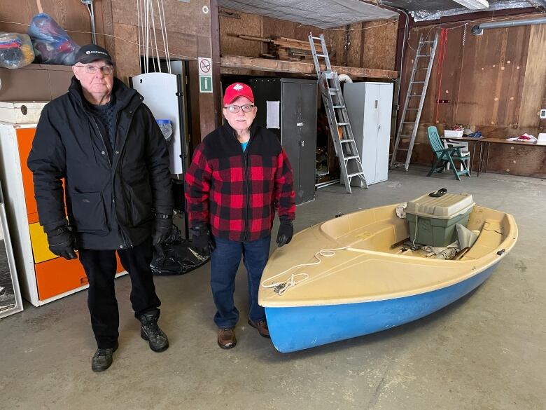 Two men stand in a rustic boathouse next to a boat on the concrete floor.