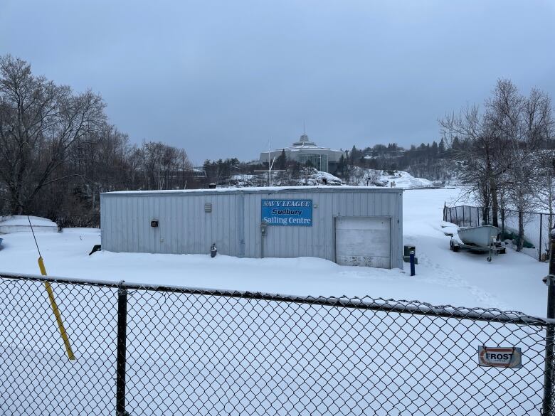 Exterior shot of a rundown blue boathouse in the winter. A science centre looms above in the background.