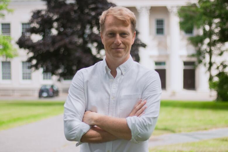 A man wearing a white dress shirt smiles with his arms crossed on a university campus.