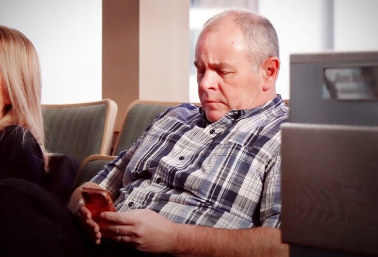 A man in a blue and white plaid shirt looks down at his phone.