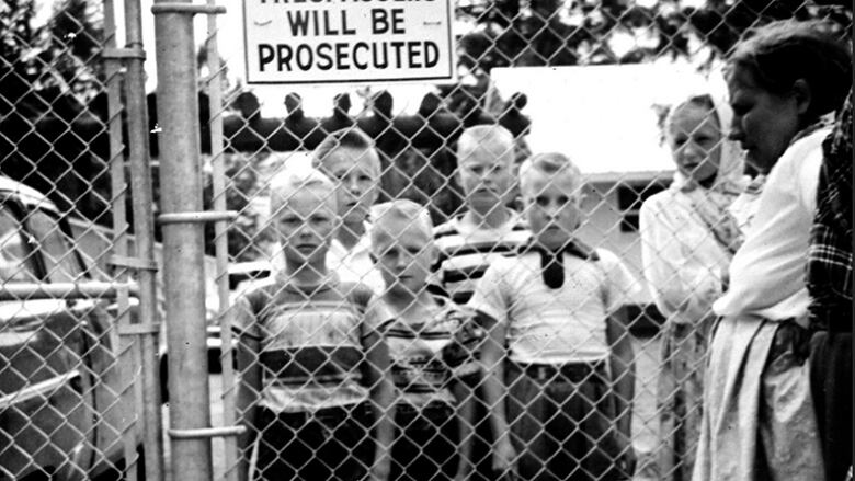 A black-and-white photo of children behind a fence with a sign reading 'Trespassers will be prosecuted.'