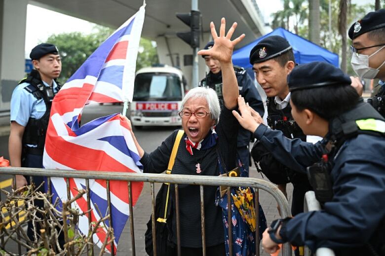 A woman with grey hair stands behind a steel barricade, holding a flag and raising her left arm. She is surrounded by police officers.