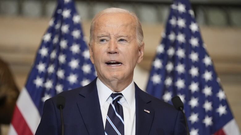 A clean-shaven older man in a suit and tie speaks at a podium in front of American flags.