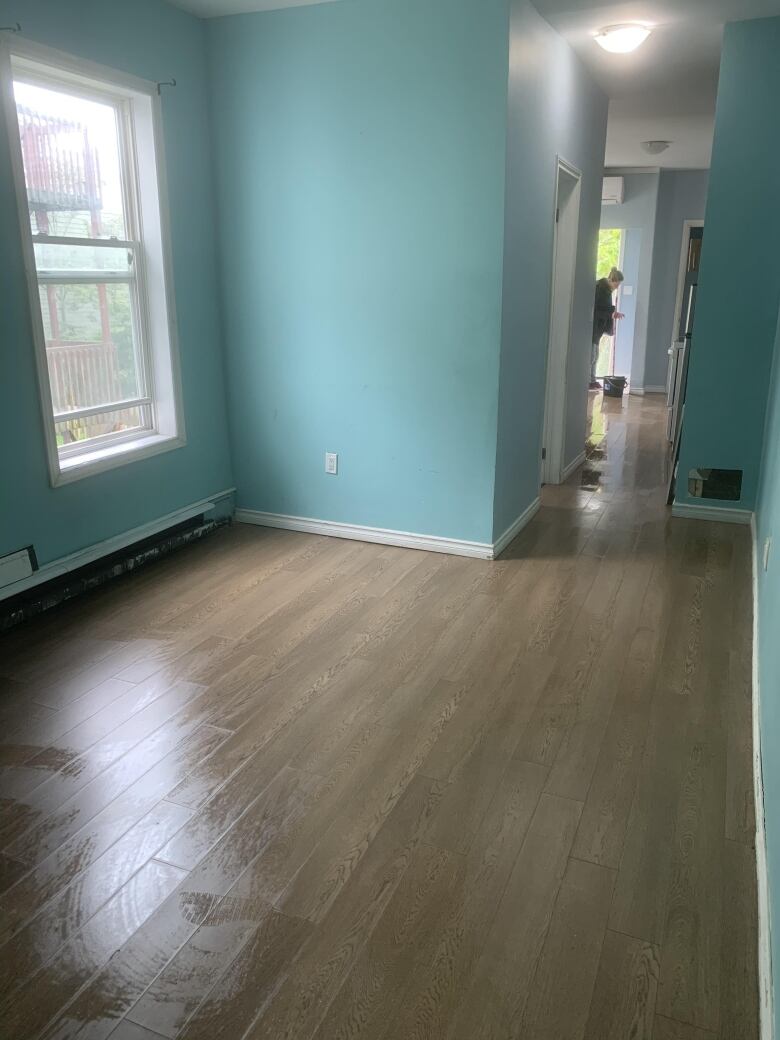 An empty room with turquoise painted walls and a gleaming hardwood floor. In the distance down the hallway near an open door a person can be seen using a mop and bucket.