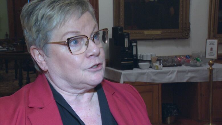 A closeup of a woman with short, blond hair and glasses, wearing a black blouse and red blazer, speaking.