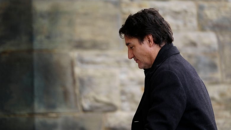 A man in a dark overcoat passes by a stone wall.