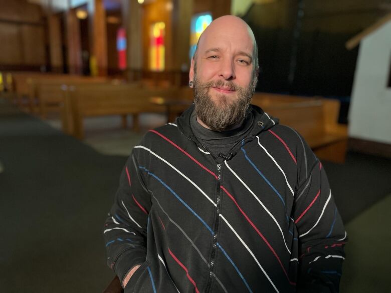 A bald man wearing a zip up hoodie stands in a church in front of colourful stained glass.