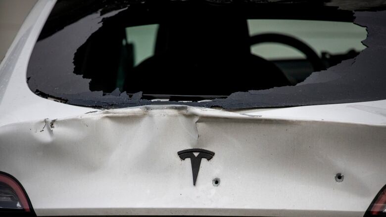 A white Tesla car's back windshield is heavily damaged and bullet holes are pictured into it.