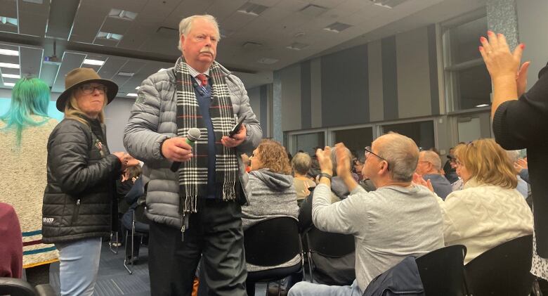 Grey-haired man with mustache, in grey puffer jacket and plaid scarf, is applauded by members of the audience as he carries a microphone. 