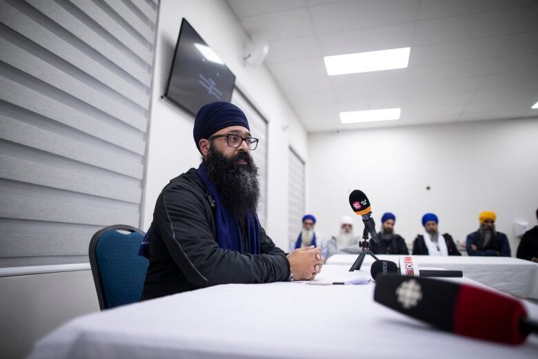 A Sikh man wearing a turban delivers a news conference while other Sikh men are behind him in the background.