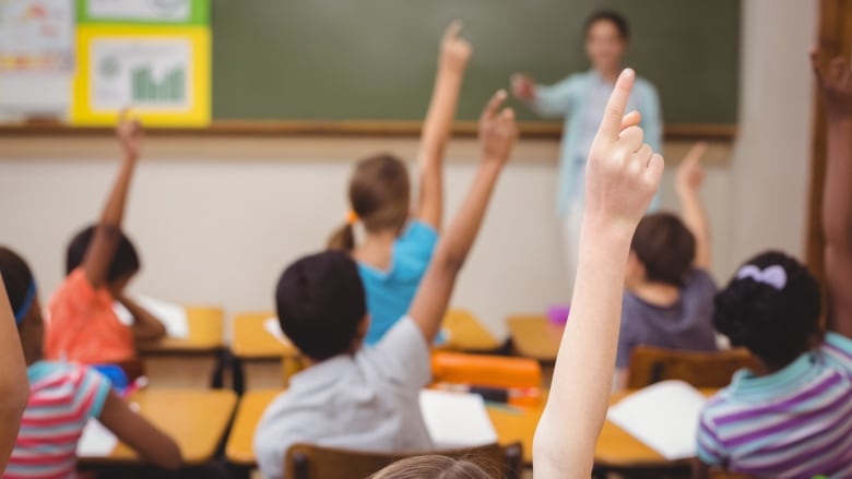 A teacher stands at the head of a classroom, with students raising their hands.