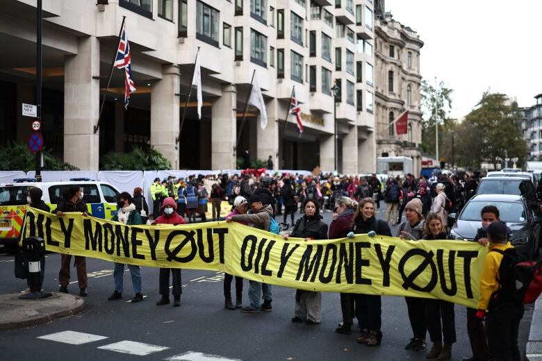 Several people are shown holding up a banner across a street that says 'Oily Money Out,' in front of a building.