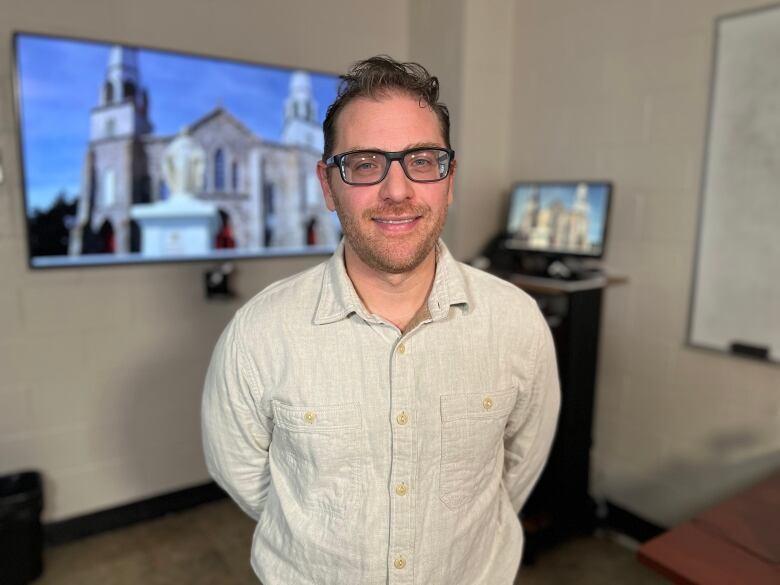 A smiling man wearing a white shirt and glasses. He's standing in front of a TV, which has a picture of an old stone church on it.