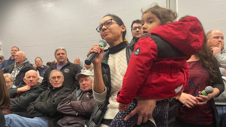 A woman in a white jacket and black-framed glasses holds a young child in her arms and speaks into a microphone.