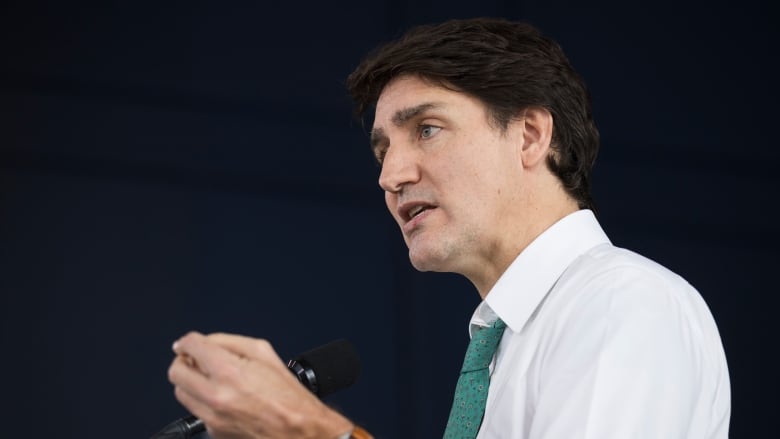 Trudeau wears a white shirt and gestures with his hand as he answers a question at the podium. 