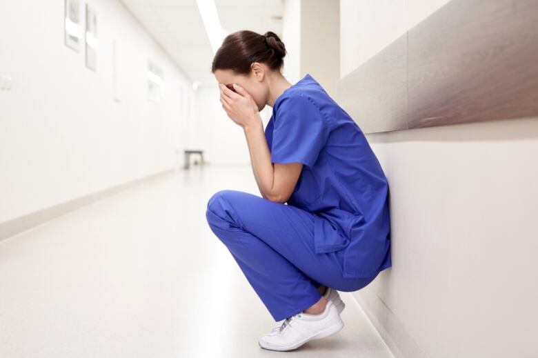 An upset nurse in blue scrubs crouches in a sterile hospital hallway.